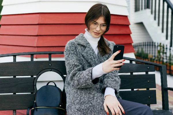 Une élégante fille aux cheveux bruns avec des lunettes et un manteau gris s'assoit sur un banc en plein air et fait du selfie — Photo