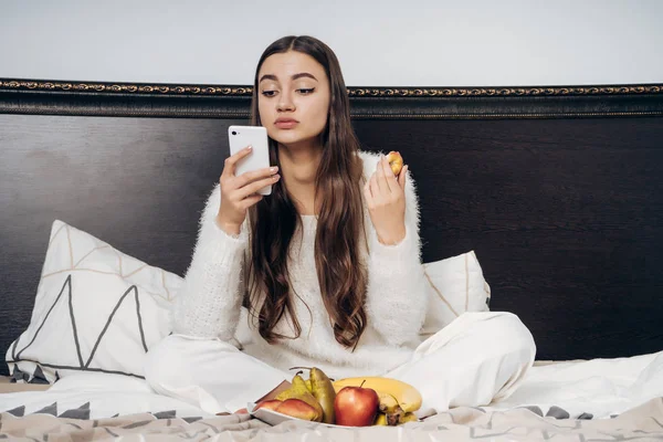 Hermosa chica de pelo largo se sienta en la cama, descansando después del trabajo, comiendo frutas y mirando en el teléfono inteligente — Foto de Stock