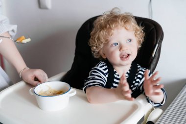 Küçük bir kıvırcık saçlı çocuk çocuk koltukta oturur, dostu aile, genç bir bakım anne bebeğini beslemeleri.