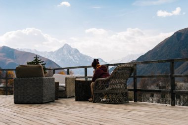 Kız gitti, ahşap bir açık veranda üzerinde oturur ve görkemli yüksek dağlar görünüyor