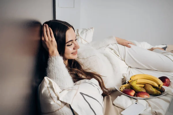 Feliz chica de pelo largo acostada en la cama, descansando después del trabajo, junto a un plato con fruta útil — Foto de Stock