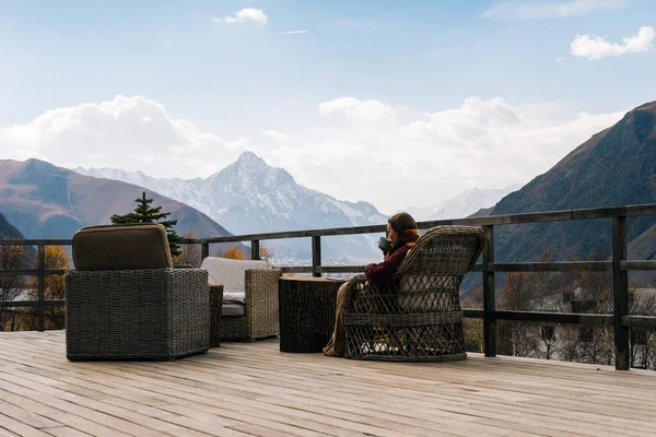 Ein junges Mädchen reist, sitzt auf einer offenen Veranda, trinkt Tee und genießt die Berglandschaft — Stockfoto