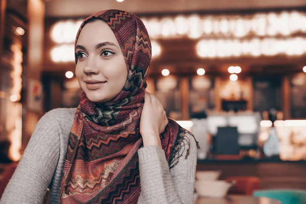 Молодая восточная девушка в хиджабе сидит в кафе, улыбаясь и глядя в окно — стоковое фото