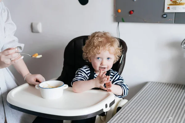 Маленький ребенок, мальчик, сидит в детском кресле на кухне, заботливая мать кормит своего сына ложкой — стоковое фото