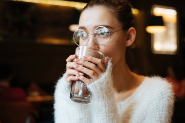 sevimli genç kız öğrenci gözlük ve beyaz bir ceket bir kafede oturan ve lezzetli bir kapuçino içmek