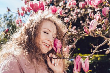 kıvırcık saçlı bir gülen genç kadın parkta yürüyor, bir güzel kokulu pembe Manolya ve sıcak hava sahiptir