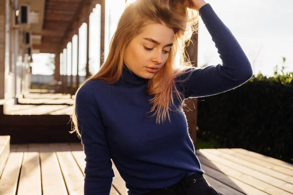 Confident girl blond model in a trendy blue sweater sits in the park posing in the sun — Stock Photo, Image