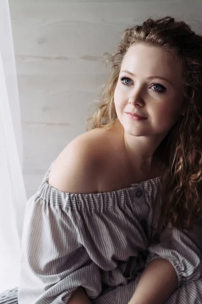 an attractive curly-haired woman in a dress sits by the window, thinks about something and smiles