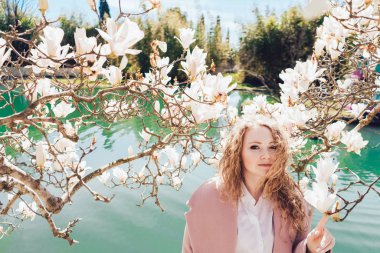 şirin, kıvırcık saçlı genç bir kadın pembe bir ceket ile bir ricola güzel kokulu bir manolya ile poz bir bahar parkı ile yürüyor