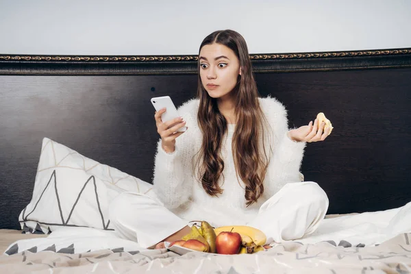 Fiatal, hosszú hajú lány ül ágyas, reggeli gyümölcs, és úgy néz ki, meglepett, az ő okostelefon — Stock Fotó