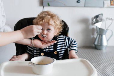 Küçük bir çocuk mutfakta bir çocuğun sandalyede oturur, sevecen bir anne oğlunu yararlı yulaf lapası ile beslenir.