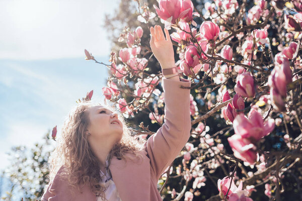 Happy curly young woman walks through the spring park, enjoys a fragrant pink magnolia