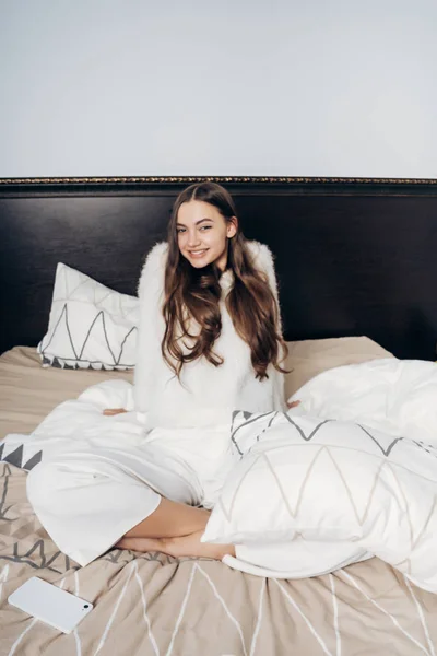 Счастливая длинноволосая молодая девушка в белой пижаме сидит в постели, просыпается утром в хорошем настроении — стоковое фото