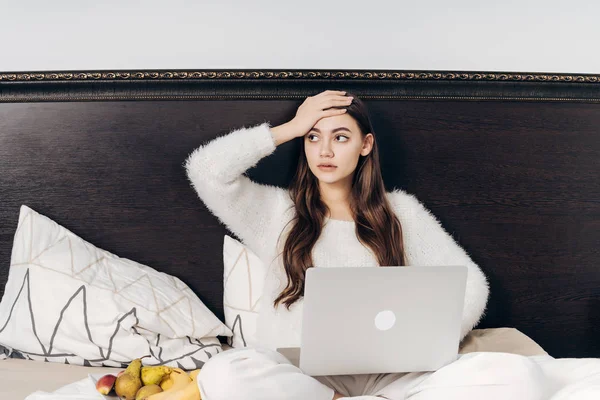 Chica joven se acuesta en la cama, viendo series de televisión en el ordenador portátil, sorprendido, junto a un plato de fruta — Foto de Stock