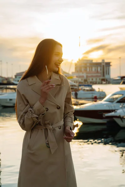 コートで笑っている髪の長い女の子のメガネを保持、夕日の光線で海によって港に立っています。 — ストック写真
