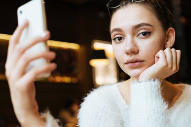 sevimli genç kız öğrenci bir beyaz ceketli okuduktan sonra bir kafede oturuyor ve bir akıllı telefon tutuyor