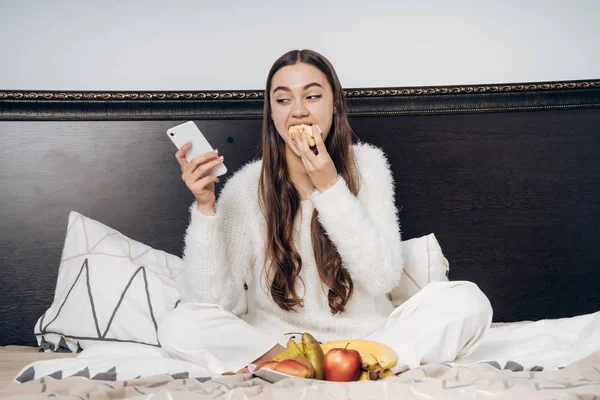 Schattig langhaar meisje in pyjama zitten in bed vroeg in de ochtend, eten van vruchten en kijken naar haar smartphone — Stockfoto