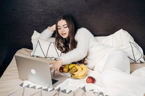 Schattig jong meisje met lang haar ligt in bed, rust, kijken naar film op haar laptop en eten van fruit — Stockfoto