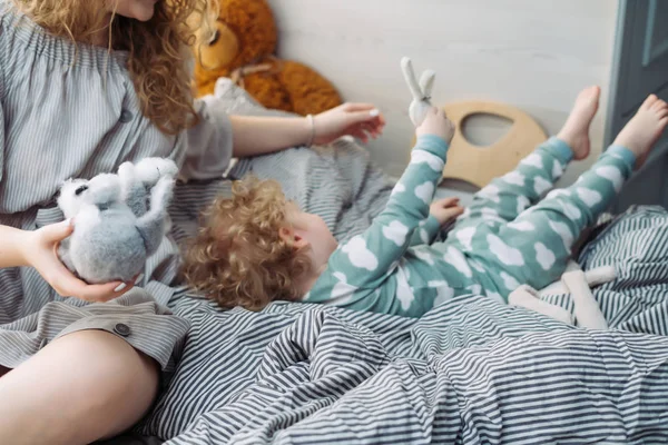 Eine junge Mutter spielt mit ihrem lockigen kleinen Kind, der Junge liegt im Schlafanzug auf einem Bett — Stockfoto