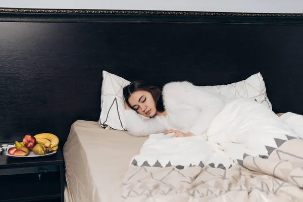Χαριτωμένο νεαρή κοπέλα σε λευκό πιτζάμες κοιμάται στο κρεβάτι νωρίς το πρωί, δεν θέλει να σηκωθεί — Φωτογραφία Αρχείου