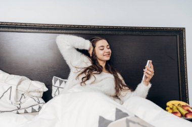 güzel uzun saçlı uykulu kız pijama erken uyanır, yatağında yatıyor, bir smartphone ve gülümsüyor tutar