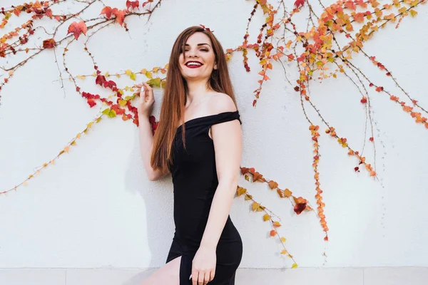 Розкішна довгошерста дівчина в чорній сукні позує на стіну і посміхається — стокове фото