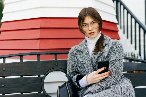 Fille élégante aux cheveux bruns dans un manteau gris et des lunettes s'assoit sur un banc à l'extérieur et tient un smartphone — Photo