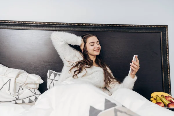 Mooie langharige slaperig meisje in pyjama vroeg wakker, ligt in bed, bezit van een smartphone en glimlacht — Stockfoto