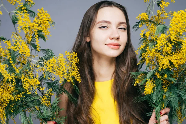 Atractiva chica de pelo largo en vestido amarillo sostiene una mimosa amarilla fragante, sonriendo — Foto de Stock