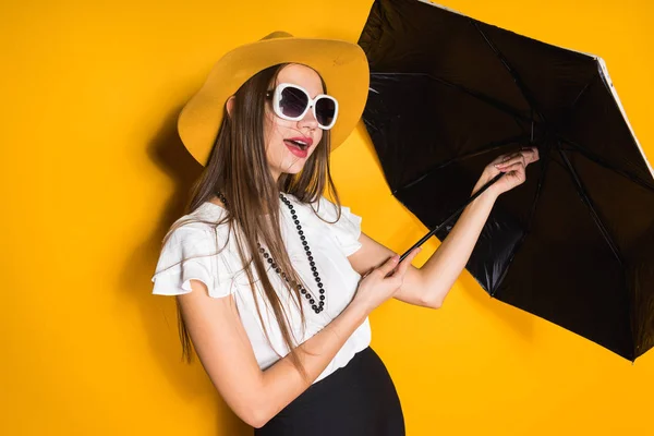 Уверенная длинноволосая модель в модной шляпе держит зонтик и позирует на желтом фоне — стоковое фото