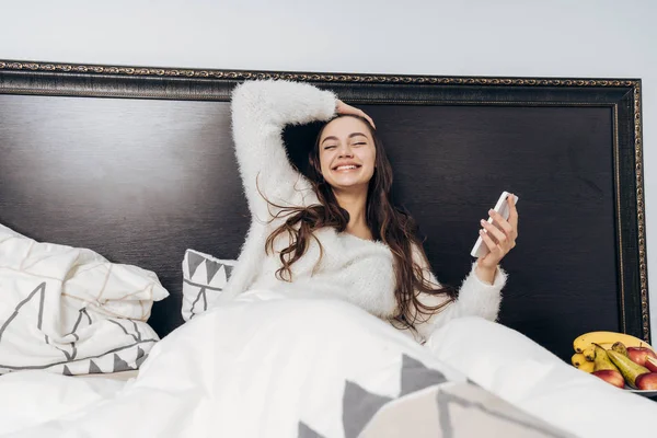 Feliz chica de pelo largo se encuentra en la cama, se regocija por la mañana, sostiene el teléfono inteligente — Foto de Stock