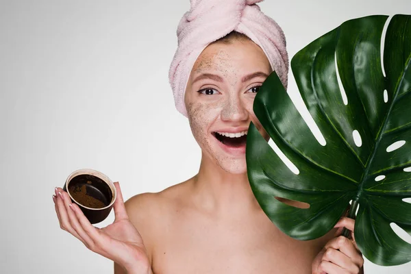 Happy νεαρό κορίτσι με μια πετσέτα στο κεφάλι της εφαρμόζεται ένα scrub στο πρόσωπό της, κατέχει ένα πράσινο φύλλο, ιαματικά — Φωτογραφία Αρχείου