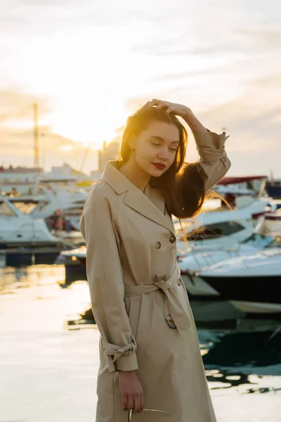 Счастливая длинноволосая девушка в стильном бежевом пальто стоит в порту на закате, улыбаясь — стоковое фото