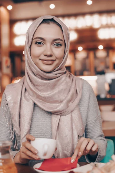 Linda garota árabe com lenço de cabeça sorrindo, sentado em café acolhedor e beber café — Fotografia de Stock