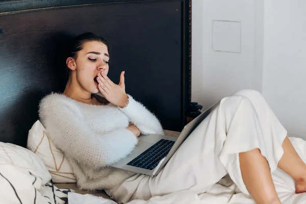 Niña soñolienta en pijama sentada en la cama, viendo series de televisión en la computadora portátil tarde en la noche y bostezando —  Fotos de Stock