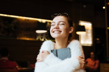 mutlu genç kız freelancer beyaz kazaklı onun defter ve gülümsüyor hugs, bir kafede oturuyor