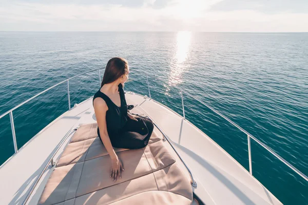 Роскошная молодая девушка в черной летней одежде сидит на белой яхте и смотрит на спокойное синее море — стоковое фото