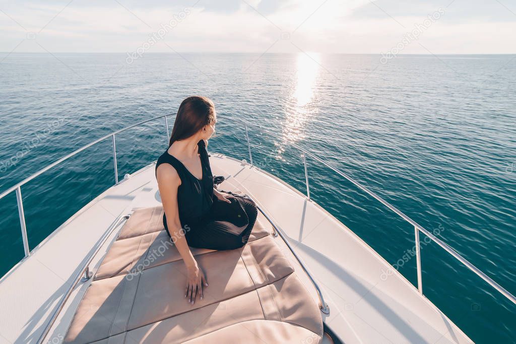 a luxurious young girl in black summer clothes sits on her white yacht and looks at the calm blue sea