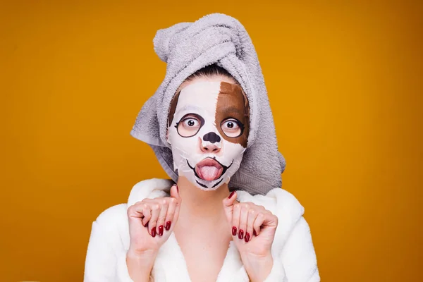 Divertida joven con una toalla en la cabeza y con un abrigo blanco muestra su lengua, en su cara una máscara con la cara de un perro — Foto de Stock