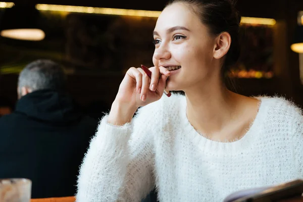 Счастливая молодая девушка-фрилансер в белой куртке сидит в кафе, смотрит в окно и думает о работе — стоковое фото