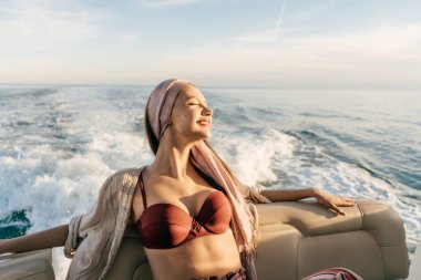 Kafasındaki bir başörtüsü ile mutlu zengin kadın akşam güneş ışınlarının onun beyaz yatta aittir, gülümsüyor ve mavi deniz üzerinde yüzüyor