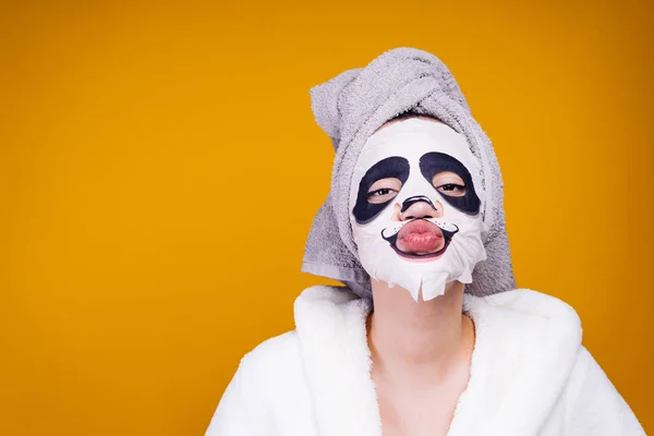 Een grappig meisje met een handdoek op haar hoofd en een witte badjas stuurt een lucht-kus, een gezichtsmasker met een gezicht van de panda — Stockfoto