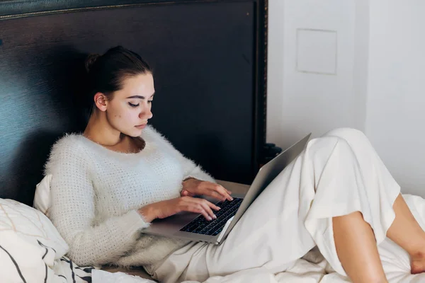 Зосереджена молода дівчина в білій піжамі сидить у ліжку і працює на своєму ноутбуці — стокове фото