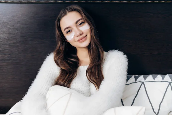 흰 잠 옷에 아름 다운 긴 머리 소녀가 앉아 그녀의 침대에서 이른 아침, 눈 아래에 흰색 패치 — 스톡 사진