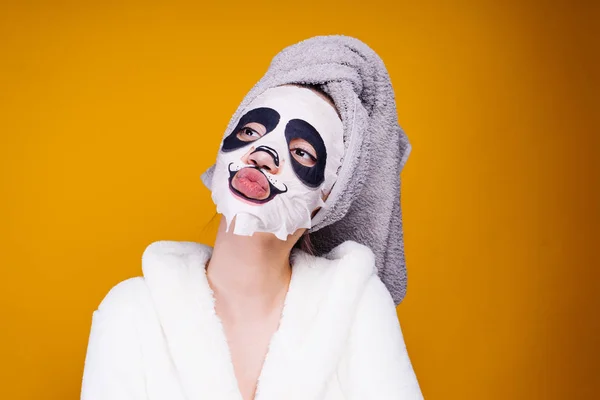 Egy vicces fiatal lány, egy törülközőt a fejét, és egy fehér fürdőköpeny, a panda arc arc-maszk — Stock Fotó