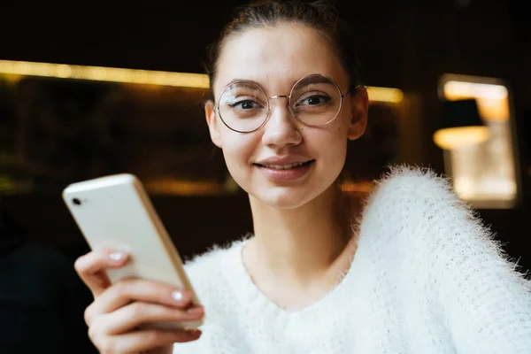 Cute młoda dziewczyna w białą kurtkę i okulary, siedząc w kawiarni po szkole, trzymając smartfon i uśmiechając się — Zdjęcie stockowe