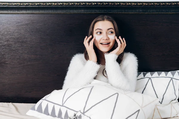 Nettes hübsches Mädchen, das frühmorgens im Bett sitzt, unter den Augen weiße Flecken gegen Schwellungen, lächelnd — Stockfoto