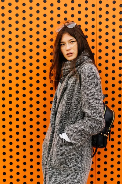 Fille de ville élégante aux cheveux foncés dans un manteau gris à la mode posant à l'extérieur d'un mur orange inhabituel en plein air — Photo