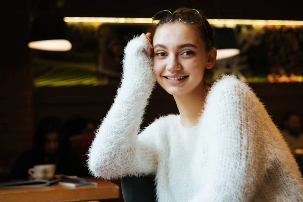 Красивая молодая девушка студентка в белом свитере сидит в кафе после учебы и улыбается — стоковое фото