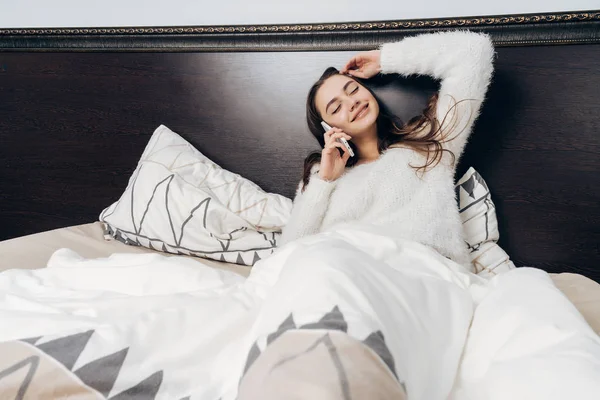 Feliz joven en pijama acostado en la cama, descansando después del día de trabajo, hablando por teléfono y sonriendo — Foto de Stock